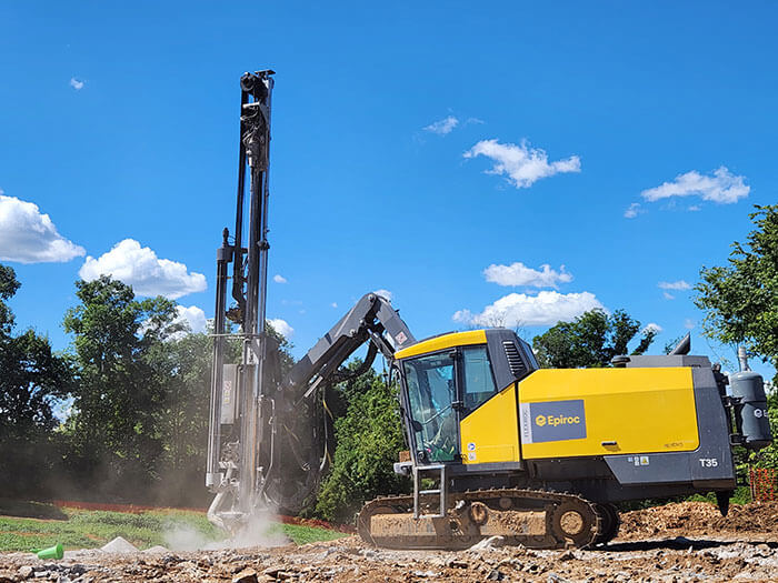 A yellow drilling machine operates on rocky terrain under a clear blue sky with scattered clouds and trees in the background, conveying a sense of industriousness.