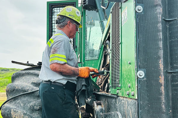 A worker in a hard hat and gloves is servicing a large green tractor outdoors. The setting is industrial, with a focus on maintenance and safety.