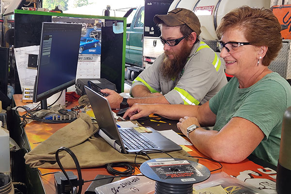 A man and woman work together in a garage. They smile while using a laptop and desktop, surrounded by tools and equipment, conveying teamwork.
