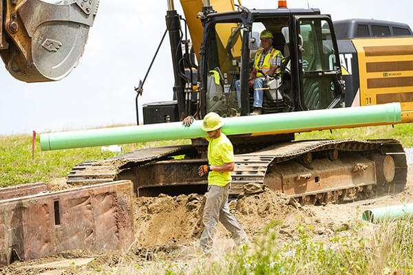 Construction site with a worker carrying a green pipe in the foreground. An excavator with a person inside is in the background on dirt terrain.