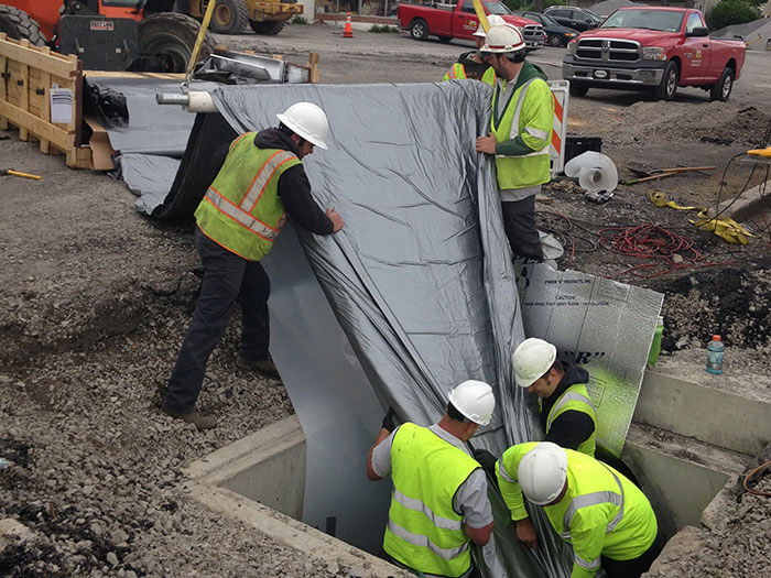 Construction workers in safety vests and helmets lay a large, reflective sheet over a concrete structure at a busy construction site. Equipment and vehicles are in the background.