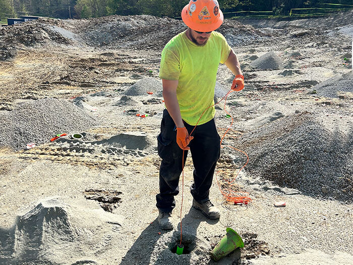 Worker in bright yellow shirt and orange helmet sets up equipment at a construction site with mounds of gravel and dirt. Sunlit, outdoor scene.