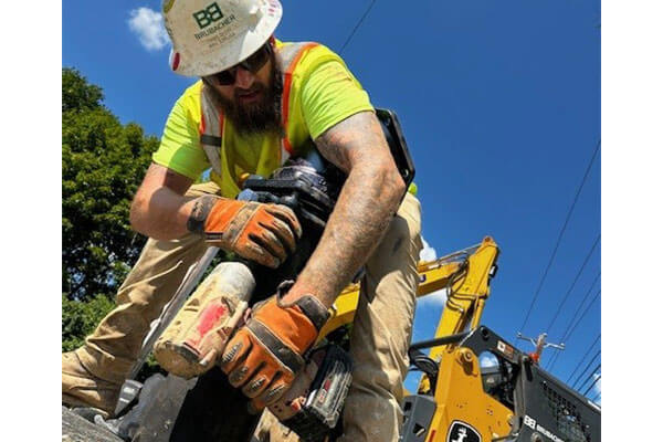 A construction worker guides a large green pipe in front of a yellow excavator on a sunny day. Another worker operates the machinery, both wearing safety gear.