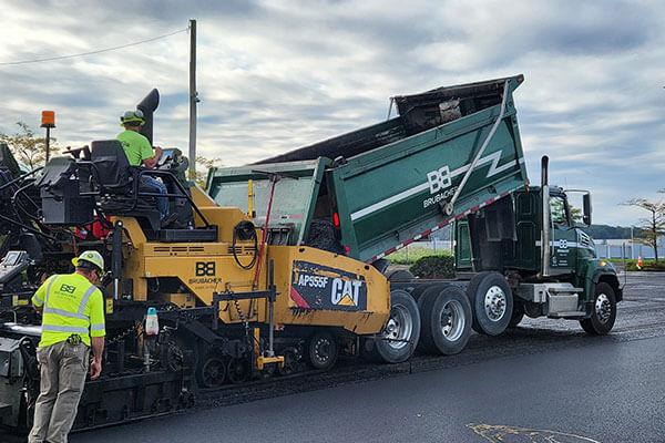 Construction workers operate a yellow CAT asphalt paver next to a green dump truck on a freshly paved road under a cloudy sky.