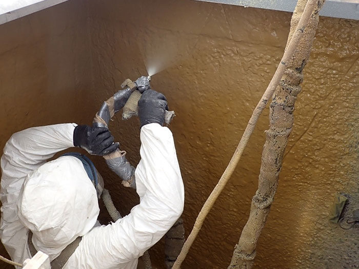 Worker in protective gear sprays sealant inside a brown, textured metal tank. Hoses and equipment surround the worker, conveying a technical environment.