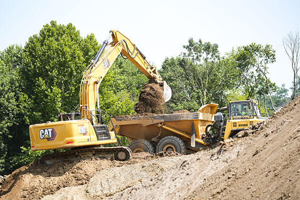 A yellow excavator loads dirt into a large dump truck on a construction site surrounded by green trees under a clear blue sky.