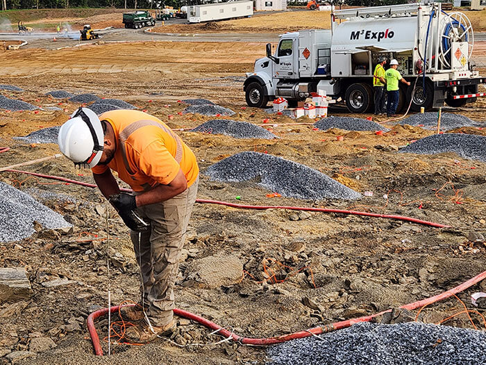 A construction worker in a hard hat and orange vest prepares a fuse on a gravel-filled site. A truck and more workers are in the background.