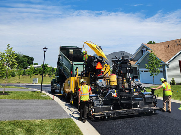Workers in safety vests operate a large asphalt paving machine on a suburban street, with a truck feeding it asphalt. The scene depicts construction under a clear blue sky, conveying a sense of industriousness and progress.