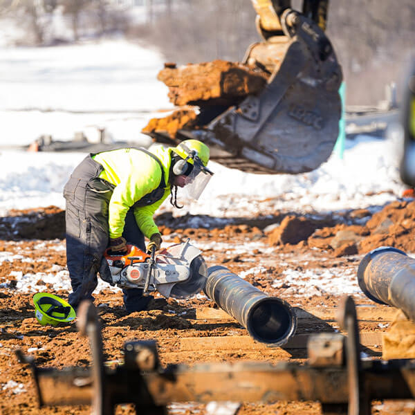 A construction worker in a bright yellow jacket uses a power saw on a large pipe at a snowy site, with an excavator in the background.