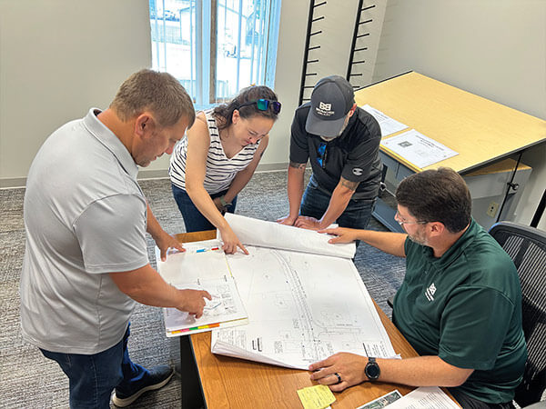 Four people in casual attire are gathered around a table, collaboratively examining large architectural blueprints. The mood is focused and engaged.