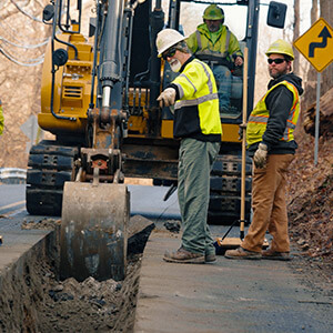 Construction workers in safety vests operate an excavator on a rural road. They dig a trench while trees and a caution sign appear in the background.