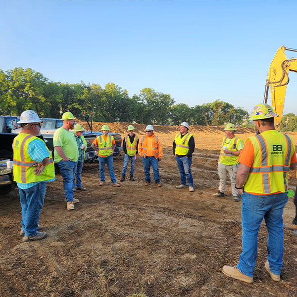 A group of construction workers in safety vests and hard hats stand in a circle on a dirt site, with machinery and trees in the background, under a clear blue sky.