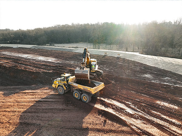 An excavator loads soil into a large yellow dump truck at a construction site with brown dirt and adjacent forest under bright sunlight.