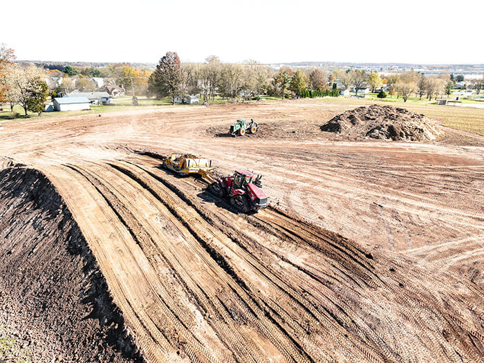 Aerial view of a construction site with heavy machinery, dirt piles, and freshly tilled earth surrounded by trees and houses.