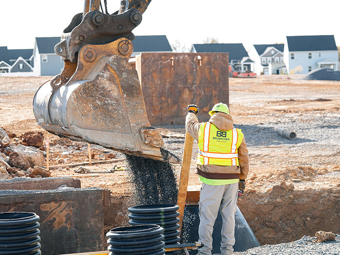 A construction worker guides dirt being unloaded from a digger into black drainage pipes on a residential site.