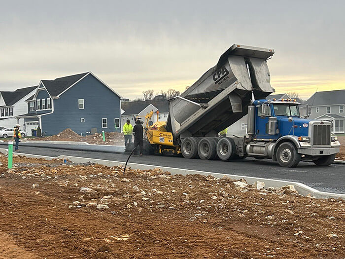 Construction crew working to pave a new road, with a dump truck unloading materials beside residential houses.