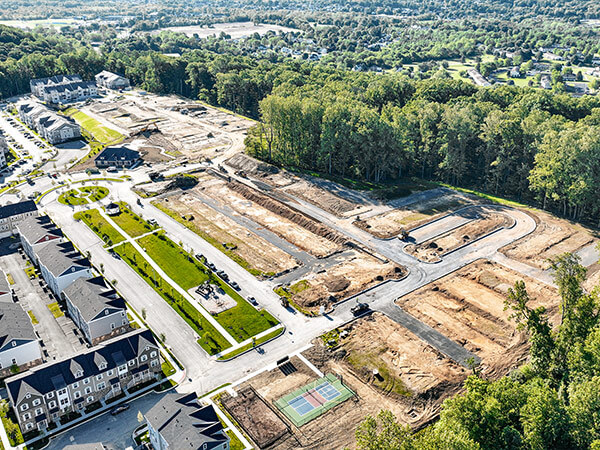 Aerial view of a housing development site with cleared plots, surrounding roads, and nearby townhouses. Green trees and a basketball court are visible.