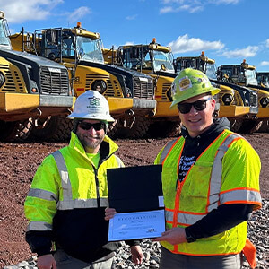 Two construction workers in safety gear stand smiling in front of a row of large yellow construction trucks. One holds a certificate under a blue sky.