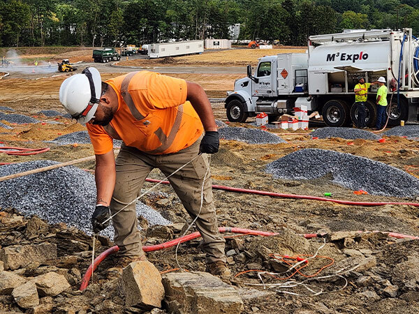 A construction worker in an orange shirt and white helmet bends over rocky ground, surrounded by gravel piles and red cables.