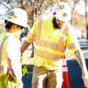 Two construction workers in yellow safety vests and hard hats collaborate on a sunny day near an orange cone, appearing focused and engaged.