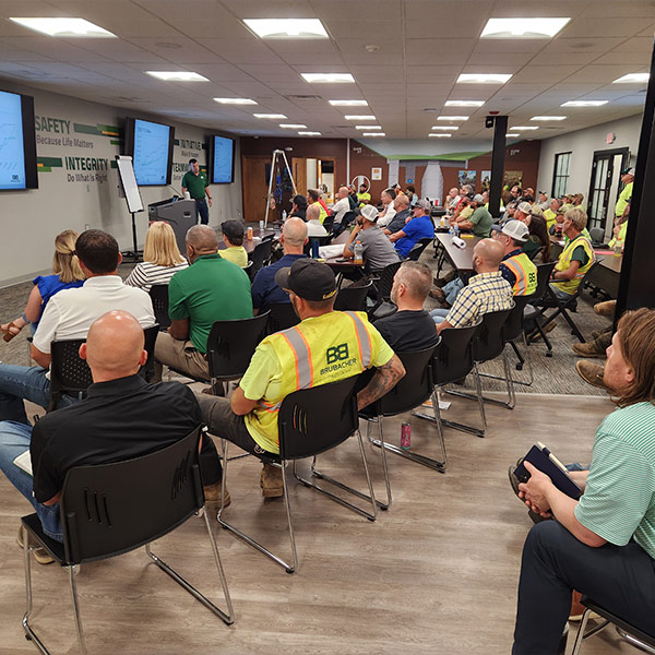 A large group of employees in safety vests sit attentively in rows during a workplace safety meeting in a conference room, with a presenter at the front.