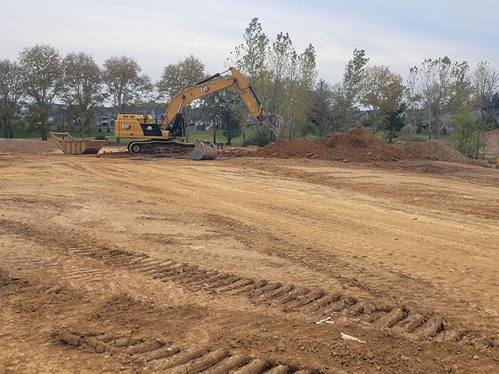A yellow excavator on a large, freshly leveled construction site, with mounds of dirt nearby and trees in the background under a cloudy sky.