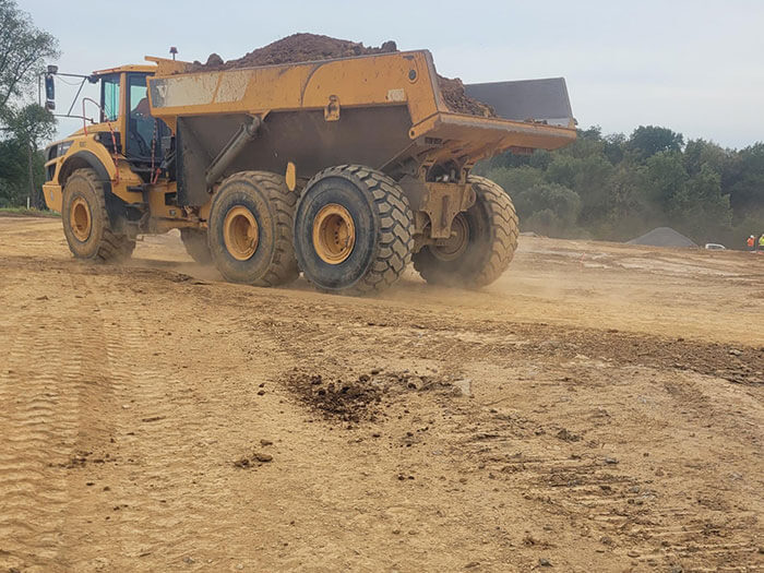 A large yellow dump truck on a dusty construction site carries a load of earth. The landscape is barren with distant trees under a cloudy sky.