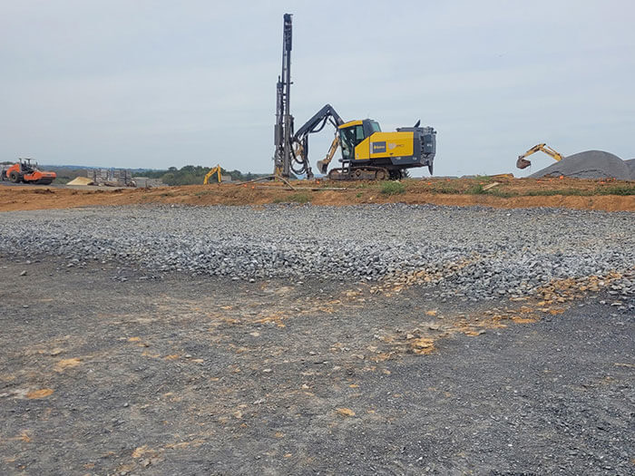 A construction site with a large yellow drilling machine positioned on a gravel surface. Additional machinery like a digger and a bulldozer are in the background under an overcast sky, conveying industrial activity.