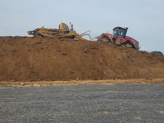 A red tractor pulling a plow is stuck on top of a large dirt mound under a cloudy sky, conveying a sense of stranded effort. Gravel ground is visible below.