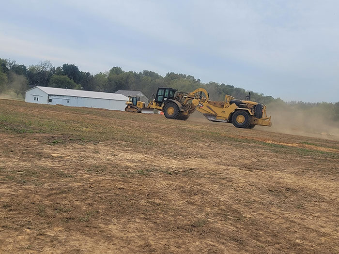 Yellow construction vehicles working on a dusty field with a white building and trees in the background, creating a sense of industrial activity.