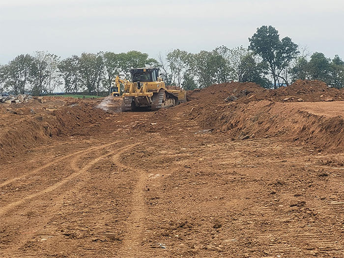 A large yellow bulldozer moves earth on a construction site, surrounded by piles of dirt. Trees are in the background under a cloudy sky.