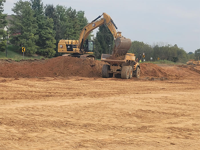 An excavator loads soil into a dump truck at a construction site, surrounded by dirt and greenery. The scene conveys industrious activity.