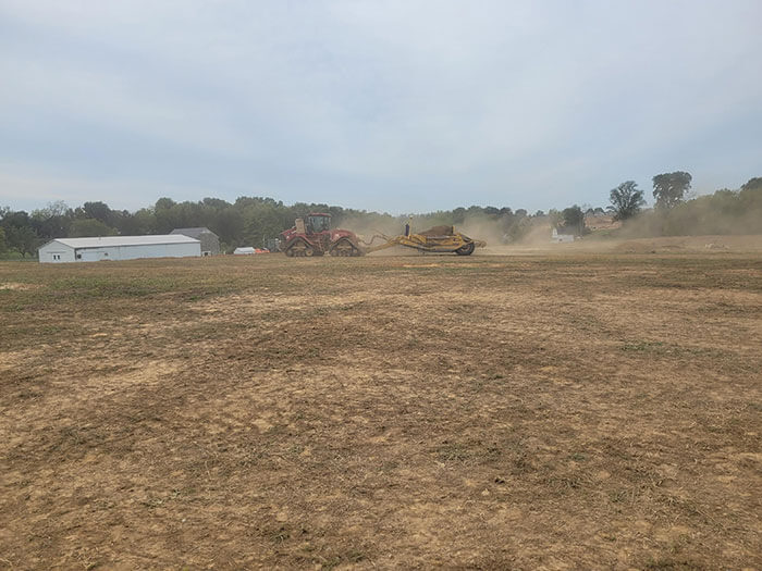 A tractor pulls a plow over a dry, dusty field under an overcast sky. In the background, a barn and trees line the horizon, creating a rural scene.